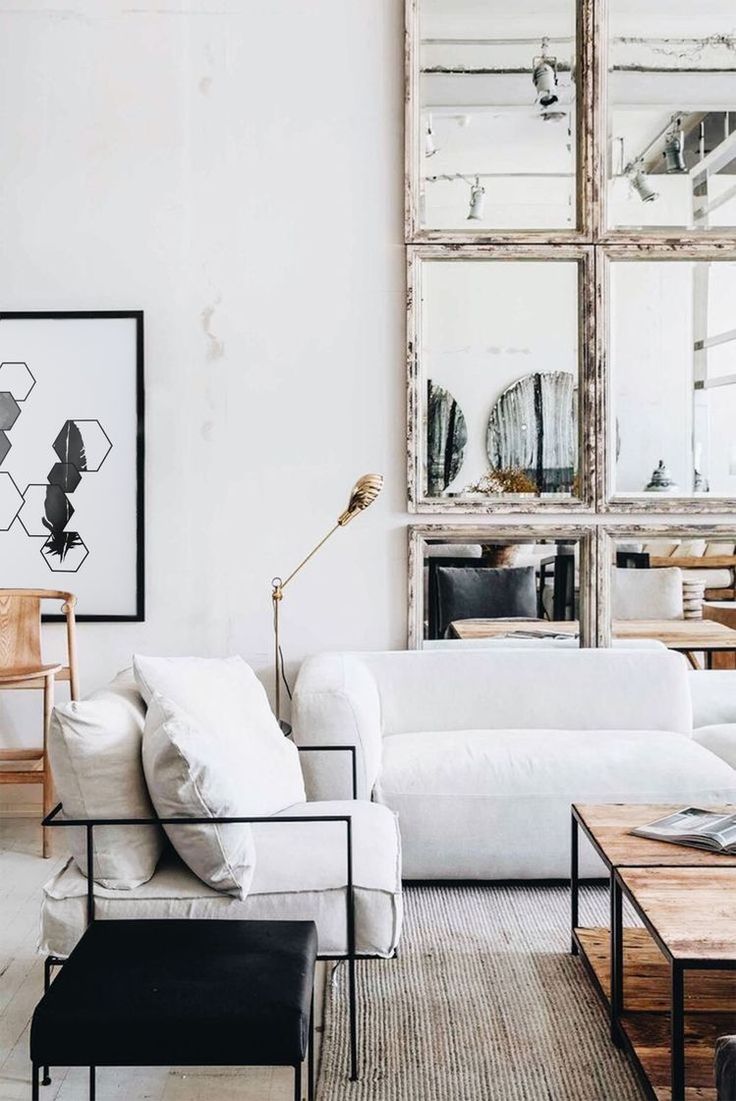 A spacious, well-lit living room with a minimalist design. Two white sofas anchor the space, facing each other. A large, multi-paneled mirror covers a significant portion of the back wall, reflecting the room and creating an illusion of greater space. A striking black and white framed print with a geometric design hangs on the left wall. A unique gold microphone-style floor lamp stands between the sofas. A dark ottoman sits in front of the sofa on the left, and a wooden coffee table with a lower shelf sits in front of the sofa on the right, displaying books and other objects. A light-colored rug with a subtle pattern lies on the floor. A portion of a wooden chair is visible on the left side of the frame. The overall aesthetic is clean, modern, and stylish.