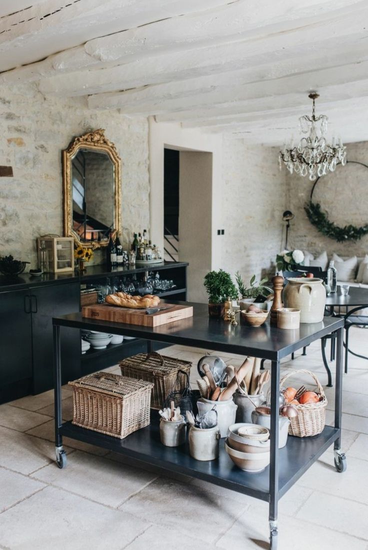 A spacious kitchen featuring a large, dark gray kitchen island with two tiers. The island holds various dishes, utensils, and decorative items, including a wooden cutting board with bread. The kitchen has textured stone walls, a white beamed ceiling, and a large gold ornate mirror. A crystal chandelier hangs above the island, and a glimpse of a living area with a wreath is visible in the background. The floor is made of large, light-colored stone tiles.