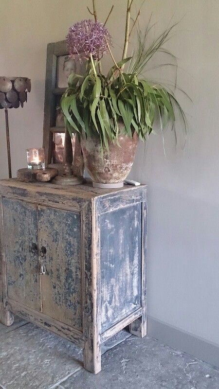 Rustic, weathered wooden cabinet in dark blue with a key in the lock, styled with a terracotta pot containing a purple allium and greenery. The arrangement includes a lamp and candle, creating a cozy and eclectic vignette.