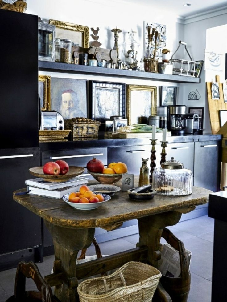 A weathered wooden table with bowls of apples, pomegranates, and oranges sits in the foreground of a kitchen with dark cabinets and open shelves filled with picture frames, figurines, baskets, and other decorative objects. A black refrigerator is visible on the left, and a coffee maker on the right.