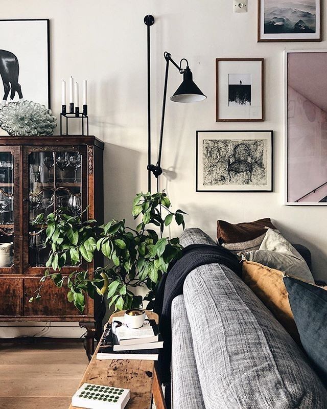 A warmly lit living room corner featuring a comfortable grey sofa with textured throws and patterned pillows. A dark wood cabinet with glass doors displays items inside to the left. Above the cabinet, a framed print with a white background and a black silhouette hangs on the wall. A collection of framed pictures and prints of varying sizes and styles are arranged gallery-style on the cream-colored wall. A tall, adjustable black floor lamp with a cone shade arches over a healthy potted plant with leafy green foliage, which sits on a small wooden table next to the sofa. A stack of books and a small white rectangular object with circular perforations rest on the table. The overall aesthetic is eclectic and inviting.