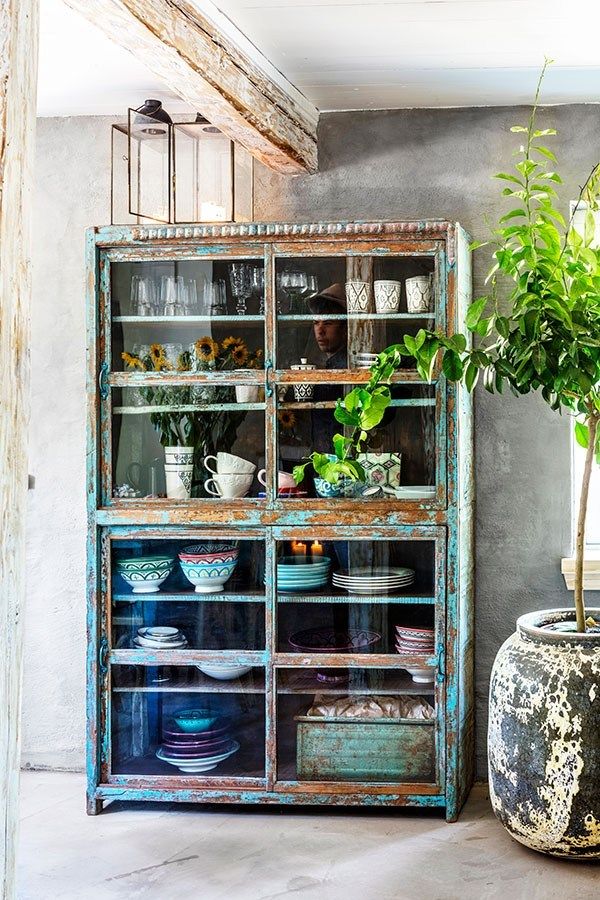 A rustic, distressed blue vintage cabinet with glass doors, filled with dishes and glassware, is placed against a gray wall with exposed wooden beams. A potted plant sits to the right.
