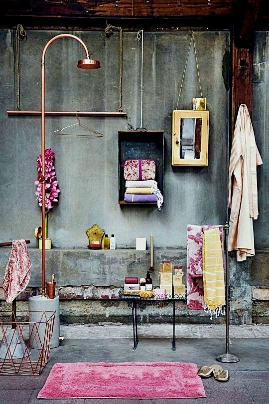 An outdoor shower setup with a curved copper pipe and rose gold showerhead against a weathered gray wall. The scene includes a hanging metal box with folded towels, a yellow cabinet with a mirror, a hanging white robe, a towel rack with patterned towels, a small table with toiletries, a pink rug, and a wire basket with a towel.