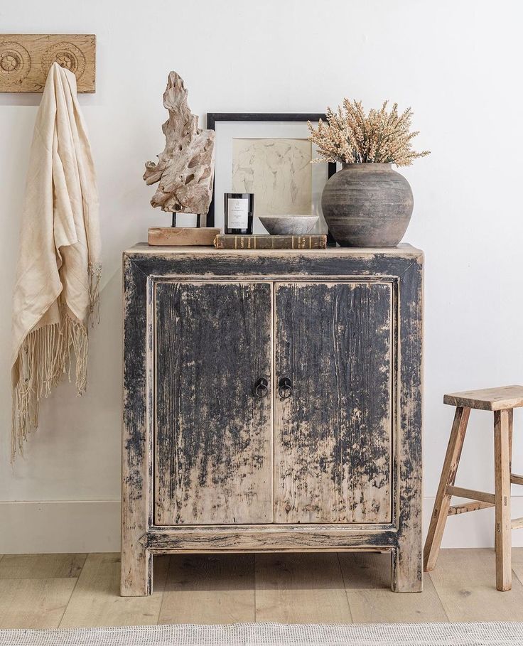A distressed two-door accent cabinet in a neutral living room, styled with a vase of dried flowers, books, a candle, and a driftwood sculpture.
