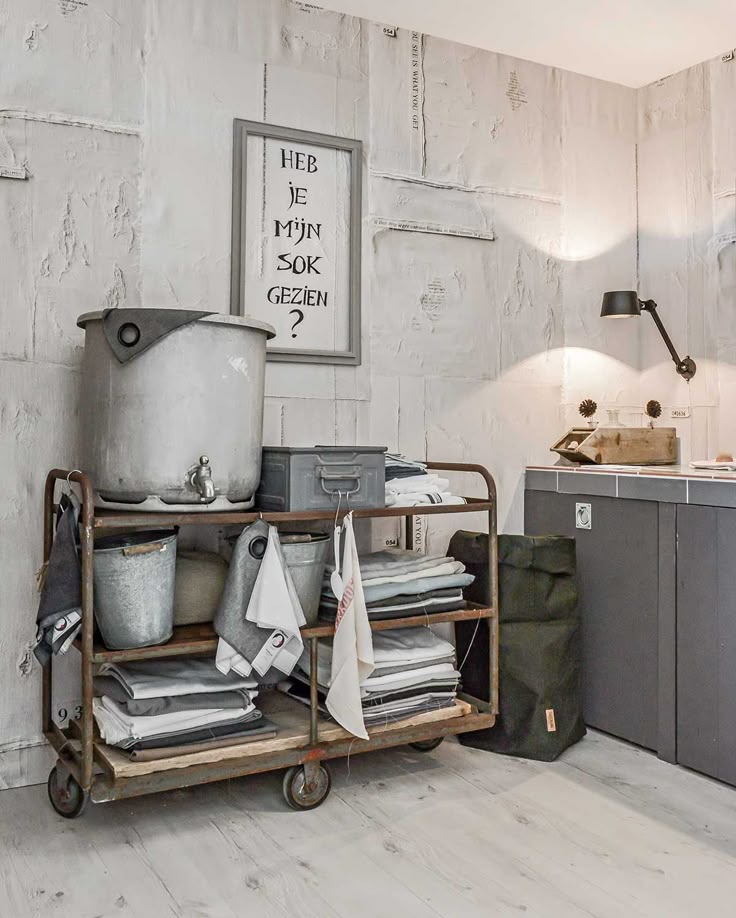 A modern laundry room featuring a metal industrial cart with wooden shelves holding a large metal container with a spigot, laundry detergent, and folded towels. The room has gray textured wallpaper, gray shaker-style cabinets, and a framed art piece with black text on a white background that reads "HEB JE MIJN SOK GEZIEN?" A black adjustable arm lamp is mounted on the wall. Two green canvas laundry bags are on the floor.