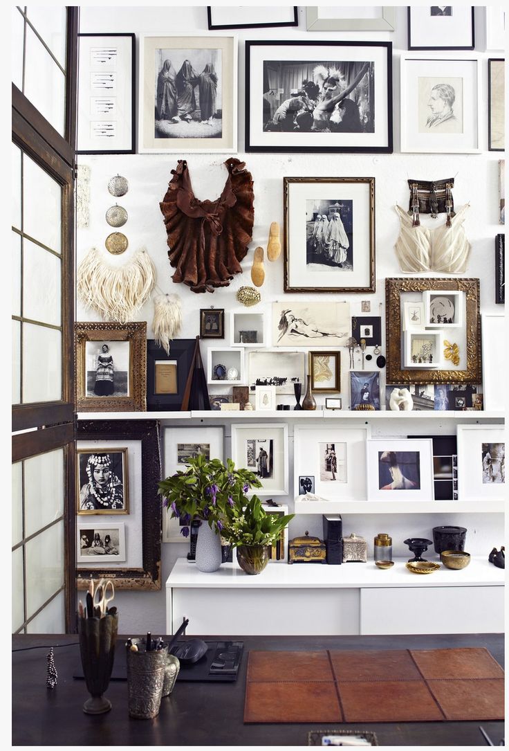 A densely packed gallery wall with a mix of framed black and white photographs, artwork, and decorative objects. The wall is behind a desk with a brown leather blotter and a vase of purple flowers. The gallery includes various frame styles and sizes, some with white mats, creating a layered and eclectic look.