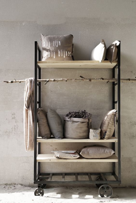A black metal rolling shelving unit with light wood shelves is styled with neutral-colored pillows, a folded throw blanket, a round woven basket containing dried lavender, and a small white textured box. The shelves are set against a textured, light-colored wall. A natural branch is hung horizontally across the middle shelf.