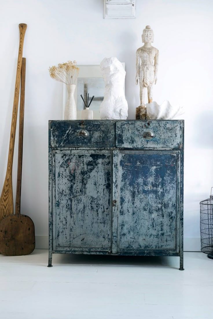 Rustic, industrial-style dark blue metal sideboard with a distressed finish, styled with eclectic decorative items against a white wall. The arrangement includes a white bust, dried flowers, a statue, and a wooden paddle and scoop leaning to the side.