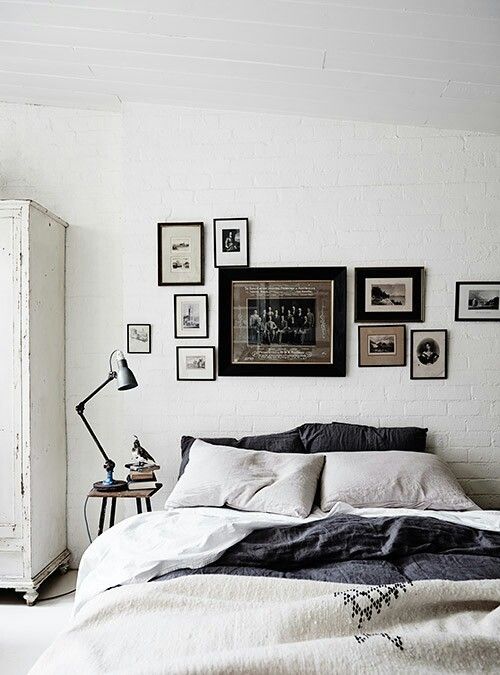 A minimalist bedroom with a white brick wall, a bed with gray and white linens, a collection of framed black and white photos arranged above the headboard, and a small side table with a black desk lamp.