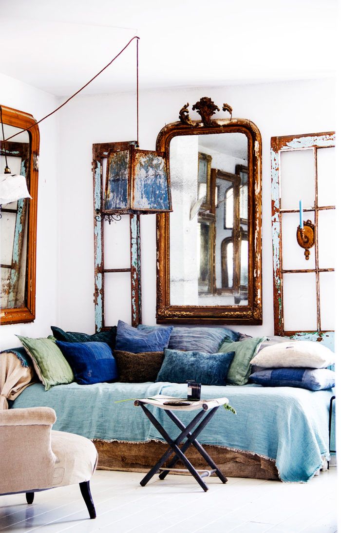 Interior with three vintage mirrors, including a large ornate mirror, flanked by distressed window frames, above a daybed with blue pillows and a throw.