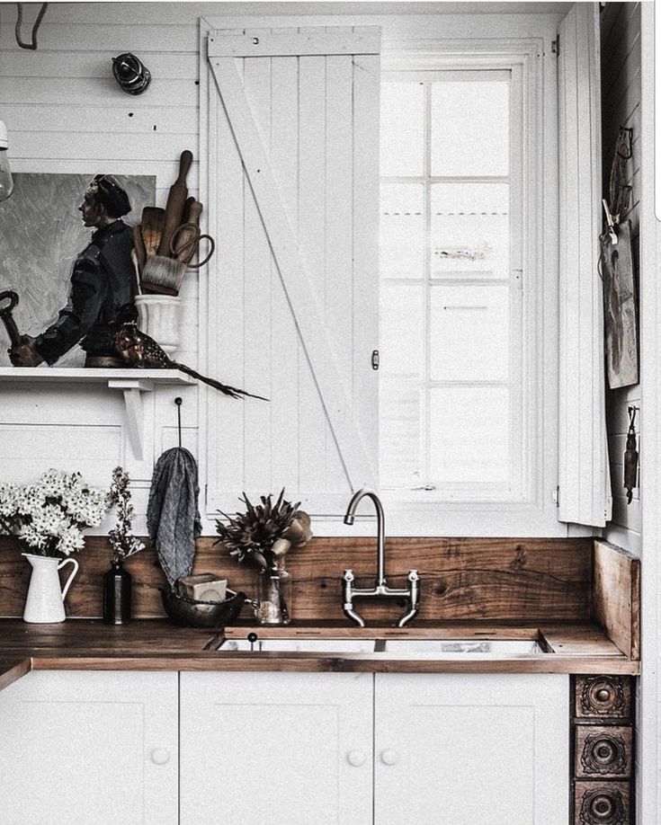 A close-up of a rustic kitchen sink area with white painted cabinets, a wooden countertop with a sink, a silver faucet, and a white shuttered window. A shelf above holds decorative items and a framed picture.
