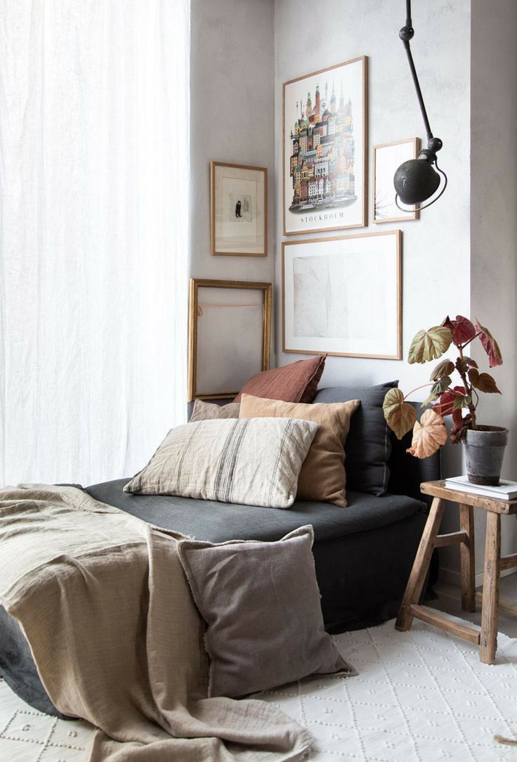 Interior decor featuring a dark gray daybed with neutral-toned pillows, framed art with a 'STOCKHOLM' poster, sheer white curtain, wooden stool with a plant, and a light geometric rug against a neutral wall.