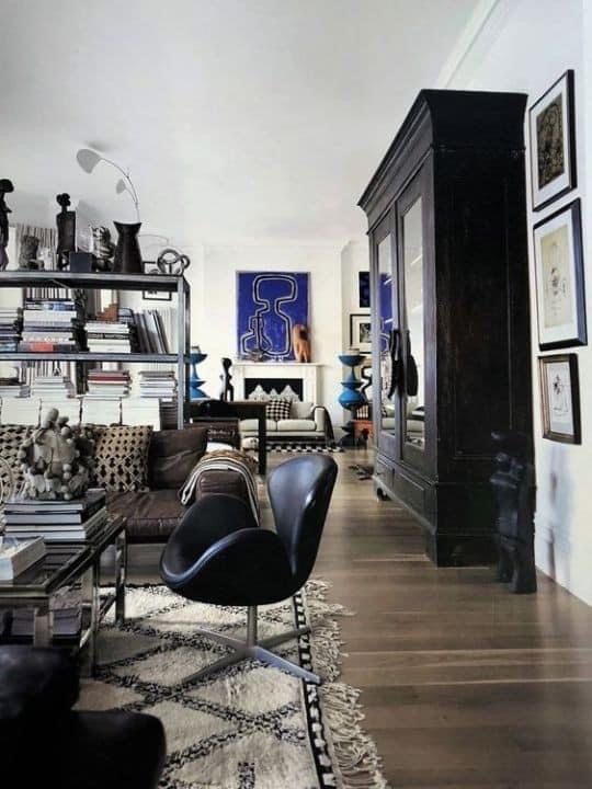 A living room with a dark, three-door bookcase against a white wall on the right. A black modern chair is in the foreground, facing a patterned rug. A metal shelving unit with books is on the left, and a sofa and fireplace are visible in the background. Framed artwork is on the wall behind the bookcase.