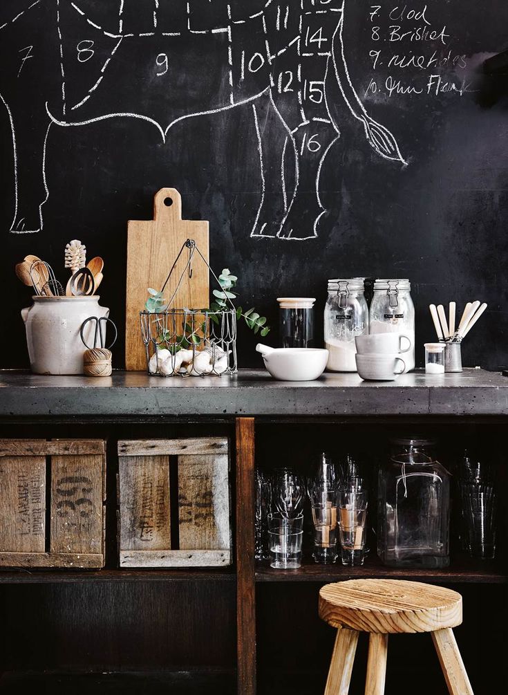 A kitchen counter featuring a cutting board, bowls, jars, and utensils, in front of a chalkboard wall with a white chalk drawing of a cow diagram with numbered sections. Below the counter are wooden crates and glass containers on a shelf. A wooden stool is in the foreground.