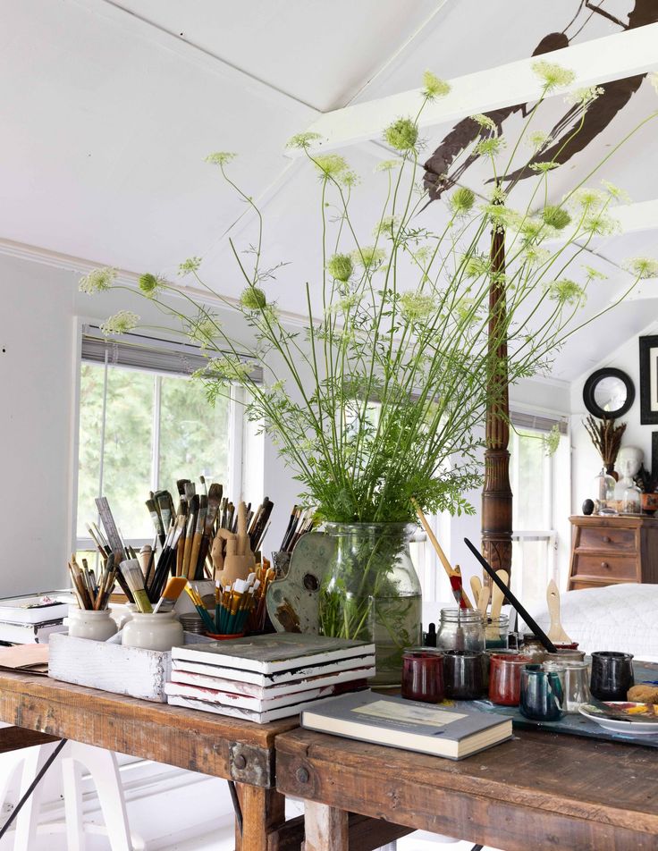 A close-up view of a rustic wooden artist's table filled with art supplies. Various paintbrushes are organized in white ceramic containers and small terracotta pots. Stacks of sketchbooks and journals are piled on the table. A large glass vase holds tall, delicate green flowering stems with white blossoms, possibly wild carrot or dill. Jars of paint in shades of red, blue, and black are arranged near the center. A small plate with food is visible on the right. The background shows a bright, airy room with a window and a glimpse of a dresser. The ceiling has a white beam with a black abstract design.