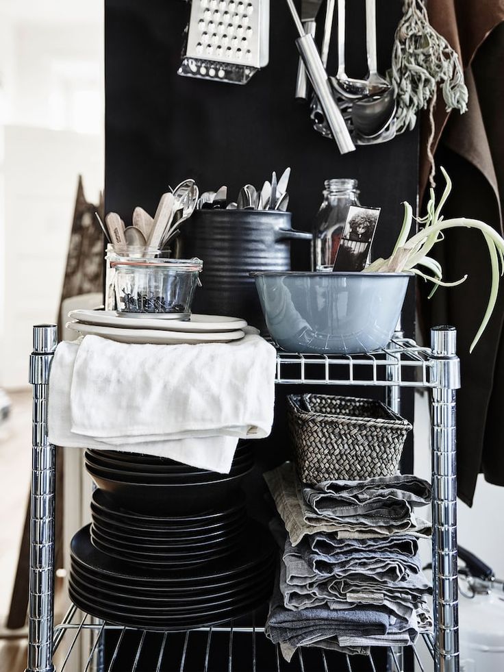 A set of chrome metal shelves filled with stacked plates, bowls, utensils, and a woven basket. A dark background features hanging kitchen tools and a plant with long leaves.