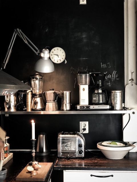 Close-up shot of a kitchen counter with a variety of stainless steel appliances, including a blender, coffee maker, and toaster. The counter is dark wood and the wall behind is a dark, possibly chalkboard, surface with the word "HEJ" in white chalk. A white clock and a silver articulated lamp are mounted on the wall.