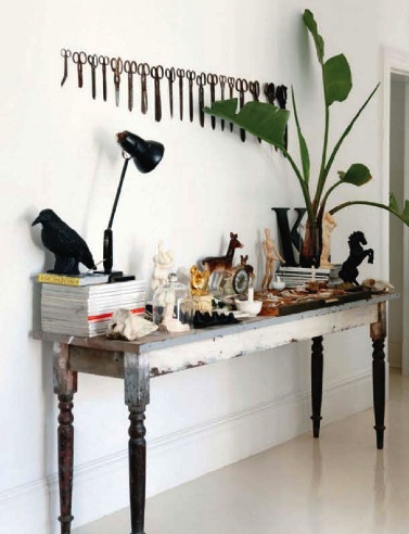 A wide shot of a rustic, distressed wooden table with dark turned legs against a plain white wall. The table is cluttered with a variety of objects, including stacks of books, a black adjustable lamp, small figurines (including a horse and a human figure), a small white bowl, and a large potted plant with broad green leaves. A black bird figurine sits on top of a stack of books. A large, dark letter 'K' is among the objects on the table. Above the table, a row of identical scissors hangs in a neat line against the white wall. The floor is a light, glossy material. The image is taken with a slightly angled perspective.