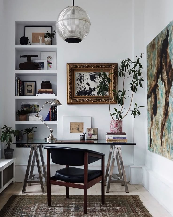A bright, minimalist home office featuring a glass-topped desk on sawhorse legs, a black mid-century modern chair, and a white wall with built-in shelves and framed artwork. A large abstract painting hangs on the right wall, and a rug is visible on the floor.