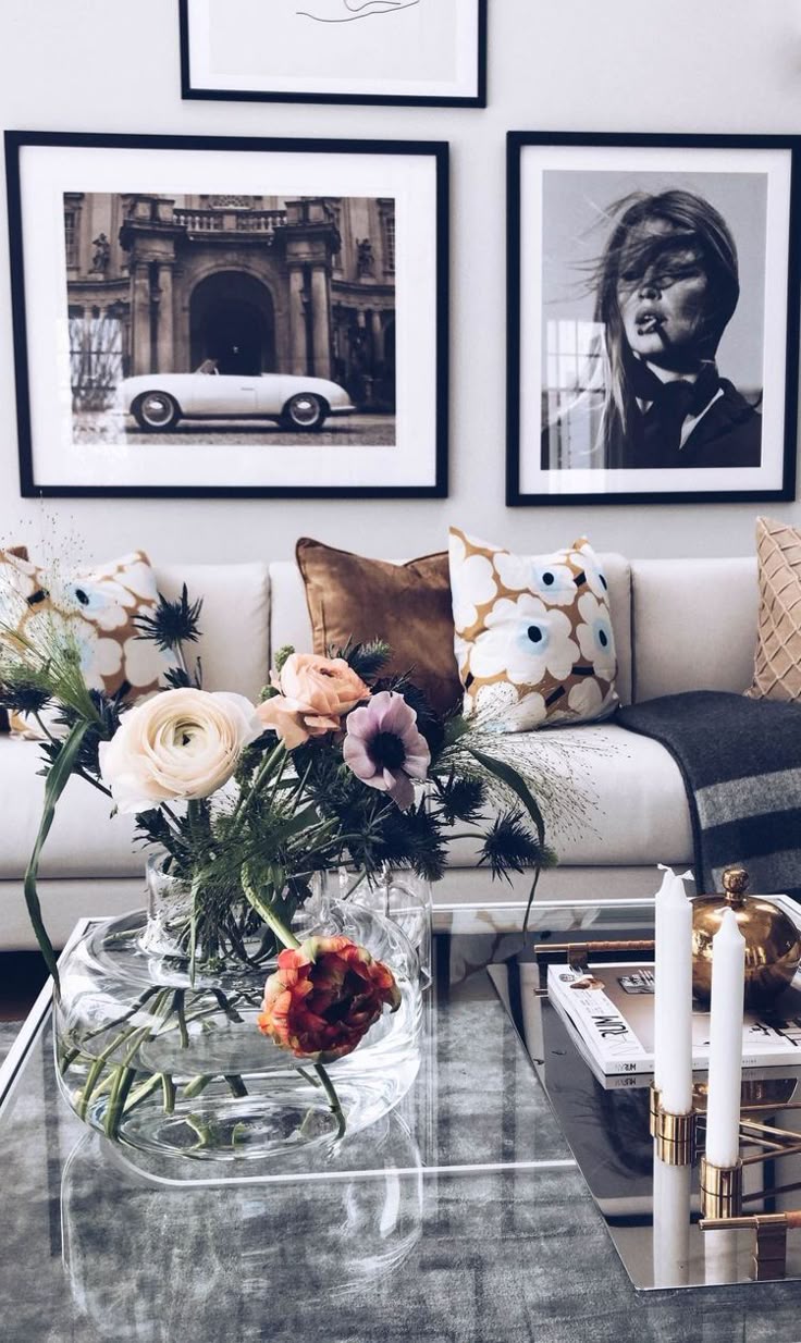 Close-up shot of a contemporary living room. A white couch with decorative pillows in floral and solid brown patterns is visible. Framed black and white photographs hang above the couch. A clear glass vase filled with pink and orange flowers sits on a glass coffee table, along with white candles and books.