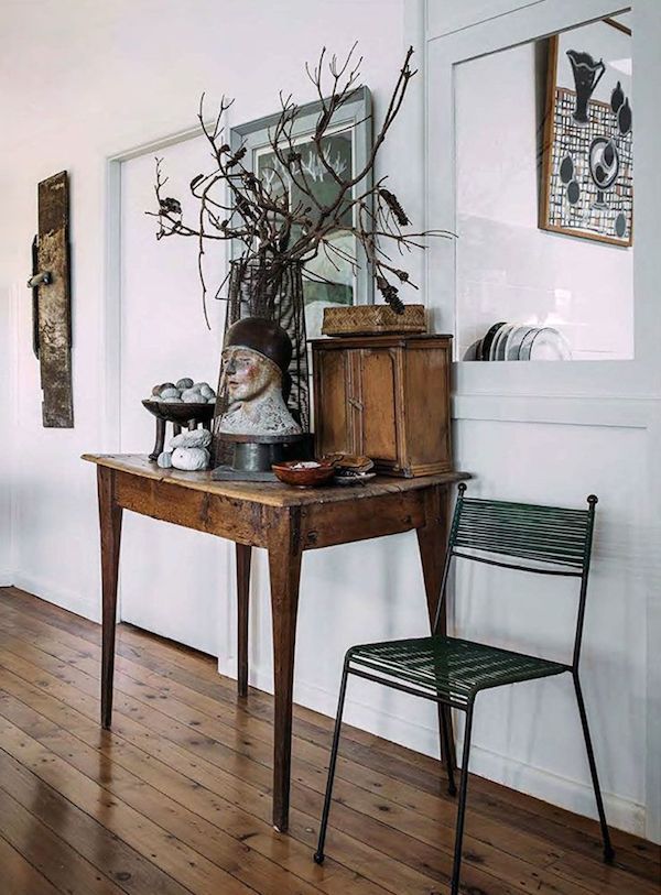 A rustic wooden table with a distressed finish stands in a room with light-colored wooden floors. On the table are a bust sculpture with a cap, a glass vase with dried branches, a small wooden cabinet, and a bowl with small stones. A green wire chair sits to the right of the table. A doorway with a white frame is visible in the background, with a framed artwork hanging on the wall to the right. A long, narrow, dark wooden object hangs on the wall to the left.