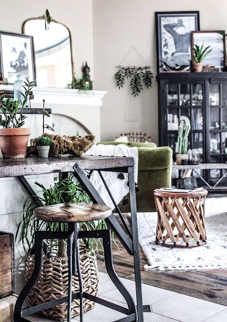 A bright and airy interior space with white walls contrasted by vibrant green plants in various pots and locations. A wooden countertop with metal supports holds several small potted plants. A tall stool with a wooden seat and black metal frame is tucked underneath, with a large green leafy plant in a woven basket beneath it. In the background, a dark cabinet displays decorative items, and more green plants are visible on top and inside. A white rug with a patterned design sits on the tiled floor.