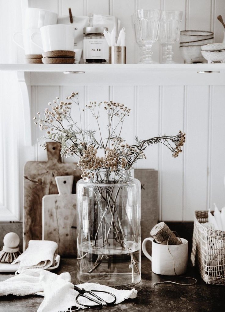 A close-up of a kitchen countertop with whitewashed paneled backsplash features a clear glass vase filled with delicate dried flowers and stems. Behind the vase are several wooden cutting boards in varying shapes and shades. To the left, a white cloth and a scrub brush rest on the dark countertop. To the right, a white mug holds twine, and a woven basket contains white items. Above, a white shelf holds white mugs, glasses, jars, and other kitchenware against the whitewashed wall.