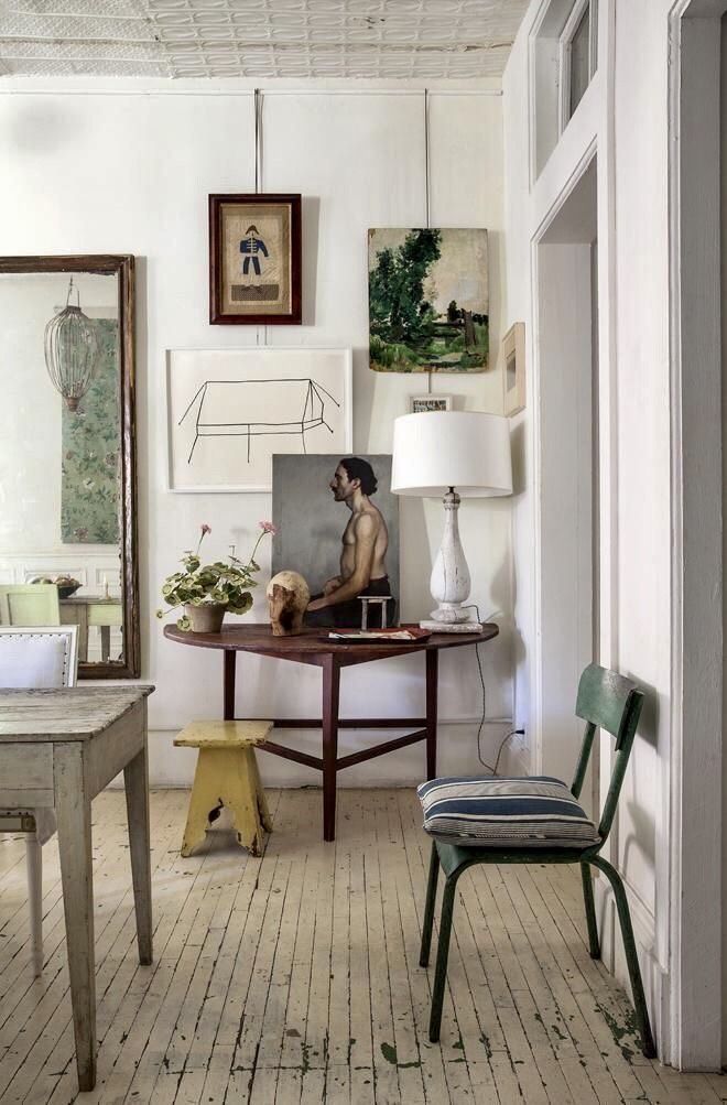 A bright and airy corner showcasing an eclectic English Cottage nook. A round wooden table holds a lamp with a white shade, artwork including a portrait and a sketch, and a potted plant. Above, various framed pieces hang on the white wall, including a folk art drawing and a small landscape painting. A small yellow stool is tucked under the table, and a green metal chair with a blue and white striped cushion sits nearby. The room features white painted wood floors and a glimpse into another room through a white doorway.