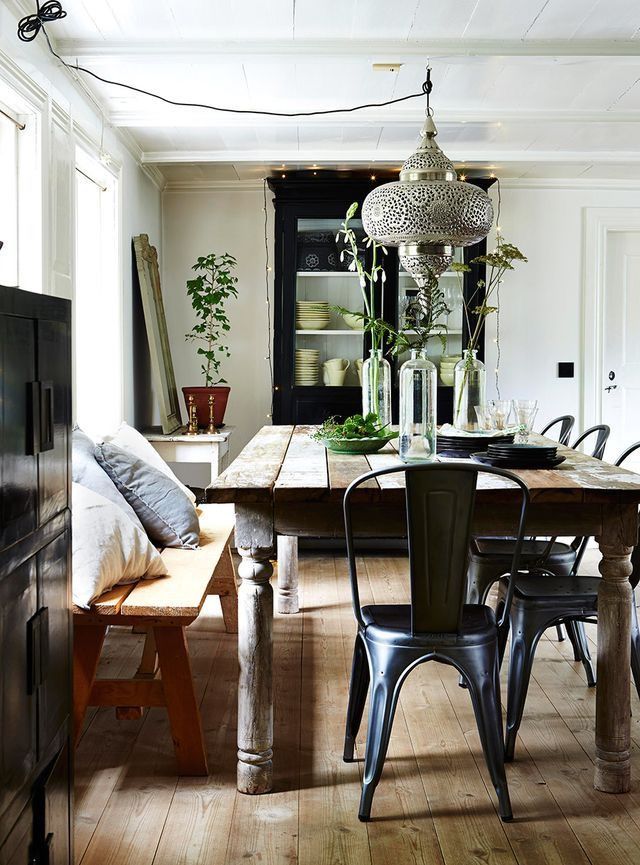 A rustic dining room with a long, distressed wooden table set with plates and glasses. Metal chairs with a vintage design surround the table, along with a wooden bench with pillows. A Moroccan-style metal pendant light hangs above the table. A black cabinet with glass doors is visible in the background, displaying white dishes. The room has light wood floors and white walls with a window. A potted plant sits on a small table near the window.