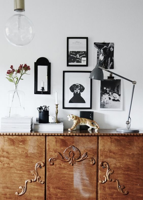 A vintage wooden sideboard with ornate carvings against a white wall. The sideboard is topped with stacks of white books, a glass vase with flowers, a black container with scissors, a white candle, and a decorative tiger figurine. A gray adjustable desk lamp stands on the right side of the sideboard. Framed black and white photos and a small black mirror are hung on the wall above. A large, clear glass lightbulb hangs from the ceiling above the sideboard.