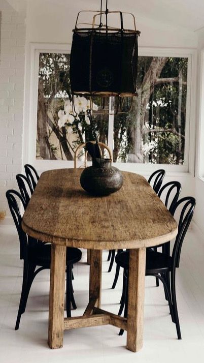 A rustic dining room featuring a weathered, oval-shaped wooden dining table, possibly a Thonet Bentwood style, surrounded by black bentwood chairs. A dark, woven pendant light hangs above the table. A dark, textured vase sits in the center of the table. A large window in the background shows trees and white flowers. The room has white walls and white flooring.