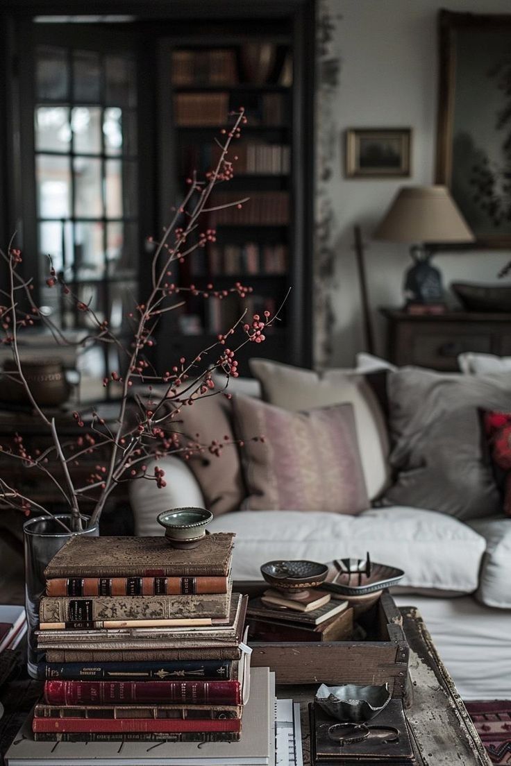 A dimly lit living room with a dark, moody aesthetic. A stack of old, worn books with colorful spines sits on a weathered wooden table in the foreground. A clear glass vase with bare branches and small red berries is placed among the books. A white sofa with assorted pillows in neutral and muted tones is partially visible in the background. A dark bookcase filled with books is seen through a multi-paned glass door behind the sofa. A vintage lamp with a fabric shade sits on a side table to the right. A small framed artwork hangs on the patterned wallpaper behind the sofa.