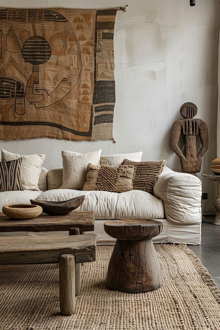 An Afro-bohemian living room featuring a light-colored, textured sofa with multiple pillows, including woven and patterned designs. A large, tapestry with tribal-inspired patterns hangs on the wall. Two rustic wooden coffee tables are in front of the sofa, one rectangular and one a round, pedestal-style. A carved wooden sculpture stands to the right of the sofa. The room has a textured, off-white wall and a woven rug with a natural fiber look.