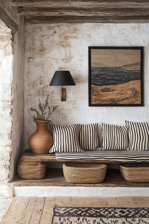 A rustic interior space with a built-in wooden bench topped with striped cushions, three woven baskets below, a terracotta vase with a sprig, a black lampshade mounted on the wall, and a framed landscape tapestry hanging above the bench. The walls are textured and whitewashed, and the floor is made of wide, light-colored wooden planks. An area rug with a geometric pattern is partially visible on the floor.