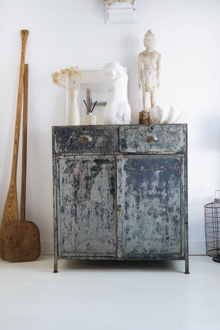 A distressed, industrial-style blue metal cabinet with two drawers and two doors is centered in the image. On the cabinet top are a white vase with dried flowers, a white abstract sculpture, a small framed picture, and a standing figure sculpture on a wooden base. A large, wooden paddle leans against the wall to the left, and a wire basket is partially visible on the right. The background is a white wall and a light-colored floor.
