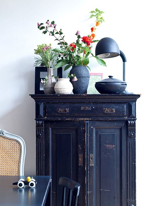 A dark blue, distressed wooden cabinet with two doors and a drawer at the top is featured. On the cabinet's surface are several vases with flowers, a black table lamp with a curved neck, and a black pot with a lid. A framed picture is partially visible behind the vases. In the foreground, a portion of a chair with a woven back and a black table with a toy car are visible. The background is a plain white wall.