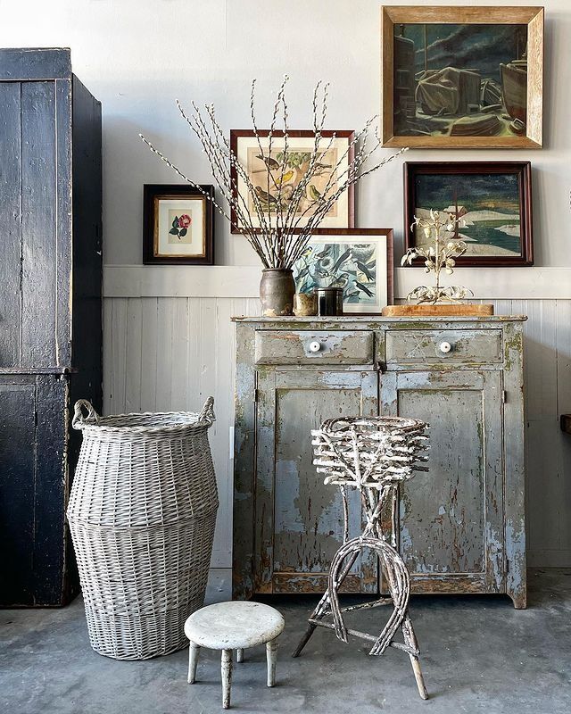 A charmingly cluttered corner showcasing eclectic English Cottage style. A weathered blue cabinet holds a brown vase with pussy willow branches, flanked by framed botanical prints and a landscape painting on a light gray wall with a white beadboard wainscoting. In front, a large woven basket, a small speckled stool, and a whimsical twig plant stand add layers of texture and interest.