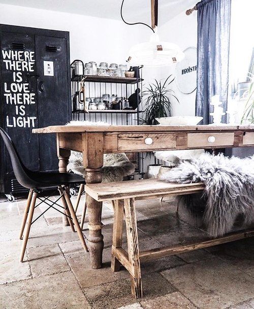 A bright dining area with textured, off-white walls features a rustic wooden table with drawers and a matching wooden bench covered with a fluffy gray sheepskin. A black modern chair with wooden legs is pulled up to the table. Behind the table, a black metal shelving unit displays various glass jars and kitchen items. To the left, a tall black locker-style cabinet has white lettering that reads "WHERE THERE IS LOVE THERE IS LIGHT". A sheer blue curtain hangs over a window, letting in natural light. The floor is tiled with light brown and gray patterned tiles.