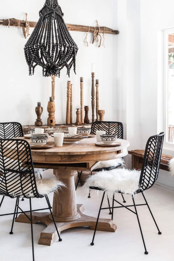 A bright, bohemian-style dining room with a round, light wood table set with plates and bowls. Four black rattan chairs with white faux fur seat cushions surround the table. A large, beaded black chandelier hangs above the table. In the background, a white wall features a rustic wooden shelf with various wooden candlesticks. A sunlit window is visible on the right.