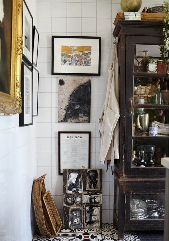 A small, eclectic bathroom corner featuring a dark, vintage cabinet with glass doors filled with various items like glass jars, metal containers, and paper goods. The cabinet stands against a white tiled wall with a series of framed artworks hung vertically. To the left, a portion of a gold ornate frame is visible. On the floor, a patterned tile design is shown, along with two woven baskets leaning against the wall. A light-colored apron hangs on the side of the cabinet.