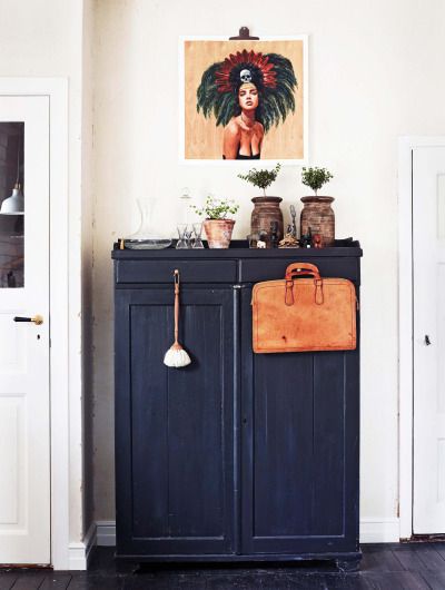 A dark blue wooden cabinet with two doors and a small drawer at the top is centered in the image. On the cabinet top are three brown terracotta pots with small green plants, a clear glass vase, and a small wooden object. A tan leather briefcase hangs on the right door, and a light-colored whisk broom hangs on the left. A framed portrait of a woman with a feathered headdress hangs above the cabinet. The floor is dark wood, and two white doors are visible on either side of the cabinet.