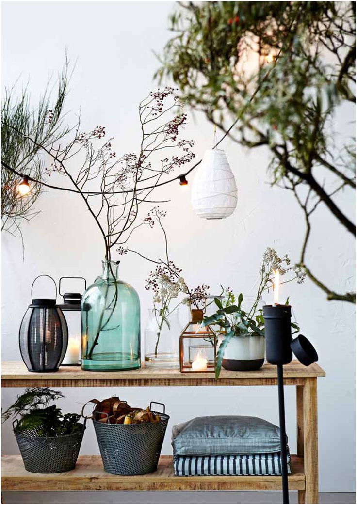 A rustic wooden shelf against a white wall displays a collection of decorative items, including glass vases with dried branches and small flowers, a black metal lantern with a lit candle, and a clear glass lantern also holding a candle. Below, two metal baskets sit on the lower shelf, one containing greenery and the other filled with brown, textured items. A folded striped textile and a gray cushion are also on the lower shelf. Above, bare tree branches and a white paper lantern hanging from a string suggest an outdoor or nature-inspired setting.