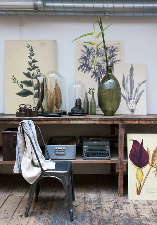 Rustic wooden console table with a collection of vintage and botanical-themed objects. The table displays framed botanical prints, glass cloches containing natural specimens, a large green glass vase with a tall flower, and a vintage typewriter. A metal chair with a draped cloth is positioned in front of the table. The floor is made of distressed wood planks, and a metal pipe runs along the ceiling.