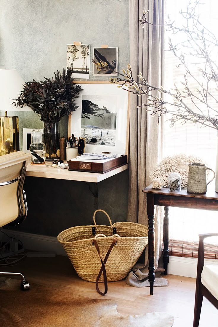 A serene home office desk vignette with a light wood desk, framed artwork, a tall vase of dried dark foliage, and vintage-style photographs pinned above. A large woven straw tote bag sits on the light wood floor with a cowhide rug accent.