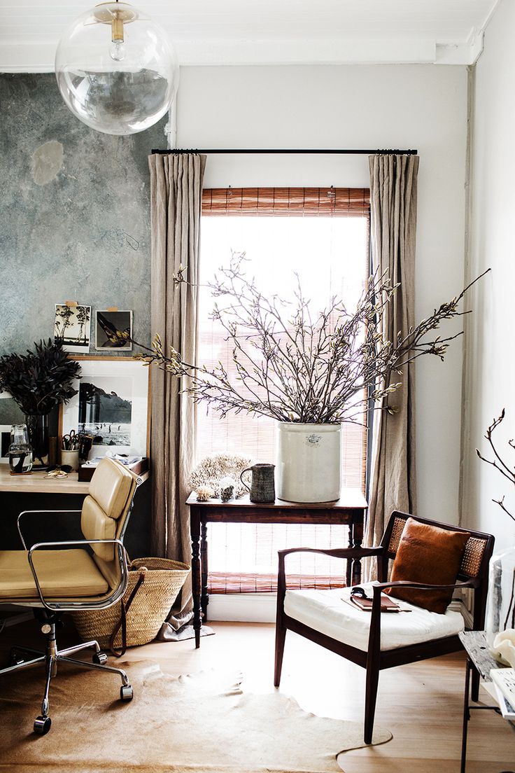 Sophisticated home office emphasizing natural textures: a rich hide rug, heavy linen drapes framing a window with bamboo blinds, and a subtly textured wall. A large ceramic pot with branches serves as a striking organic centerpiece.