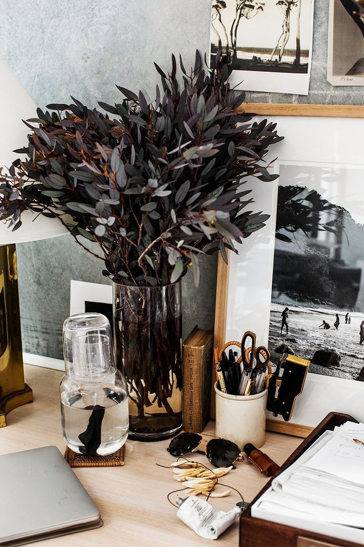 Opulent home office detail showcasing a blend of organic and refined textures: deep purple eucalyptus branches in a clear vase, framed monochrome artwork, a vintage wooden 'OUT' tray, and a brass desk lamp.