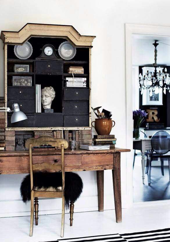 A rustic wooden desk with an attached dark hutch filled with books, plates, a clock, and a bust sits in a room with white walls and a black and white striped rug. An antique wooden chair with a black furry cushion is positioned at the desk. Through a doorway, a modern dining area with a black wall, crystal chandelier, and clear chairs is visible.