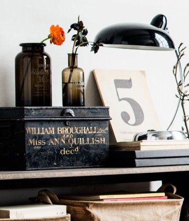 Charming cottagecore shelf display featuring natural elements like flowers in aged glass bottles, a rustic metal box, and a simple numbered print, illuminated by a black lamp.