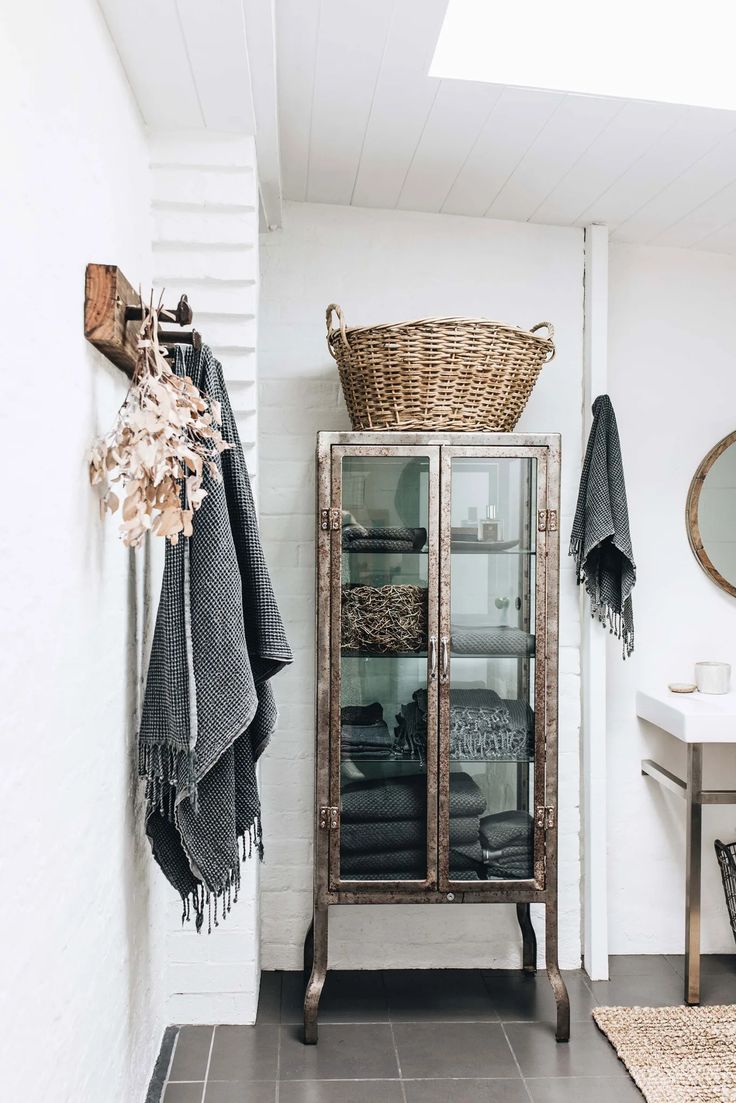Cottagecore bathroom detail featuring an antique-style metal cabinet showcasing neatly stacked gray textiles. Natural elements include a large wicker basket and dried foliage hanging on the whitewashed brick wall.