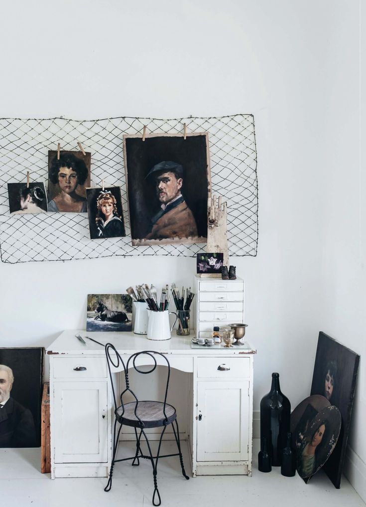 A bright artist's corner features a white vintage desk with drawers, holding paintbrushes in white pitchers, palettes, and other art supplies. Several portrait paintings are displayed on a wire grid hanging on the white wall above the desk, along with a small wooden hand model. More paintings lean against the wall on the floor. A black wire chair sits in front of the desk.